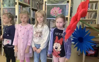 Przy regałach z książkami stoją cztery dziewczynki. Z prawej strony są sztuczne kwiaty zrobione z krepy.