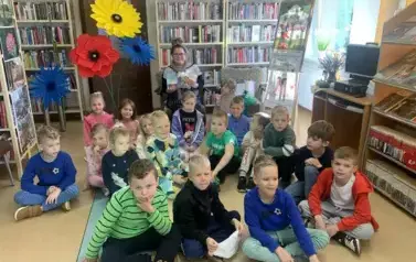 Grupa dzieci siedzi na podłodze, za nimi siedzi bibliotekarka trzymając książkę. Z lewej strony na stojaku są sztuczne kwiaty zrobione z krepy