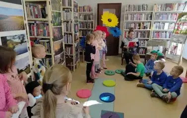 Bibliotekarka czyta bajkę,a obok stoją trzy grupy dzieci grających rolę do bajki. Grupa chłopc&oacute;w siedzi na podłodze.