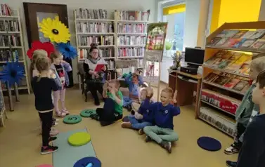 Bibliotekarka czyta bajkę,a obok stoją trzy grupy dzieci grających rolę do bajki. Grupa chłopc&oacute;w siedzi na podłodze i macha rękami.