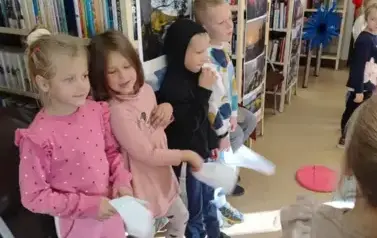 Przy regałach z lewej strony stoi czworo dzieci i machają białymi chusteczkami bawiąc się w aktor&oacute;w do " bajki o kogucie"