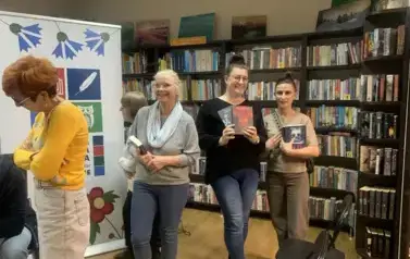 Do stolika pisarz stoją Panie w kolejce z zakupionymi książkami do podpisu. Dwie Panie  z uśmiechem pokazują swoje nowe książki.