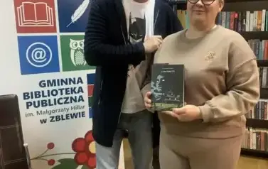 Autor stoi przy Czytelniczce, kt&oacute;ra wylosowała książkę pisarza "Czas pokuty". Trzyma książkę pokazując ją. Za nimi stoi rolap z logiem bibliteki.