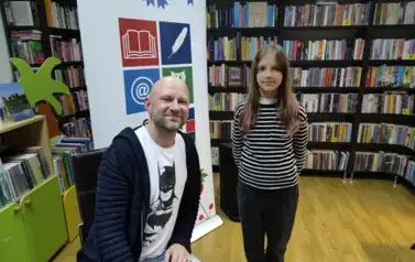 Obok pisarza stoi młoda dziewczynka dając do podpisu nową książkę. Z tyłu widać rolap z logiem biblioteki.