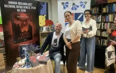 Przy stoliku pisarza stoją dwie Panie aby podpisać książkę. Uśmiechają się. Z prawej strony stoi rolap z logiem biblioteki ,a po lewej rolap promujący najnowszą książkę "Trzeci gatunek"