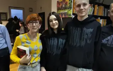Pani w rudych włosach stoi z dziewczyną i chłopakiem w czarnych bluzach. Pani w rudych włosach trzyma nową książkę pisarza. Uśmiechają się.