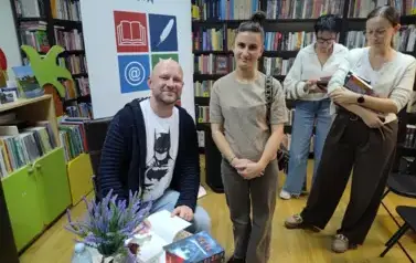 Przy stoliku Grzegorza Kopca stoją czytelnicy z nowymi książkami prosząc o podpis. Pani z przodu i pisarz uśmiechają się.