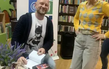 Pani w rudych włosach stoi przy stoliku pisarza prosząc o podpis w nowych książkach. Oboje się uśmiechają. Za nimi stoi rolap z logiem biblioteki.