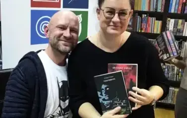 Czytelniczka trzymająca dwie nowe książki stoi blisko pisarza. Oboje się uśmiechają. Za nimi stoi rolap z logiem biblioteki.