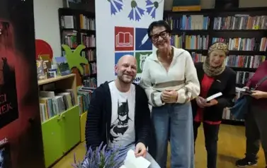 Przy stoliku gdzie siedzi pisarz stoi czytelniczka z prośbą o podpis w książce. Za Nią stoi kolejna Pani z książką w kolejce. Za nimi stoi rolap z logo biblioteki.