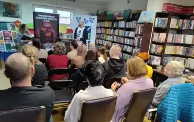 Z przodu stoją pisarz i prowadząca spotkanie. Prawie wszystkie krzesła zajęte przez Czytelnik&oacute;w.