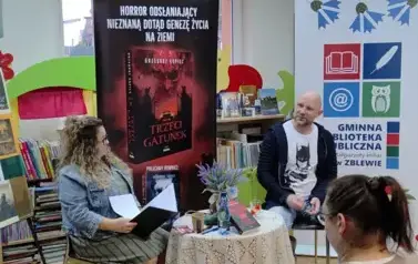 Przy stoliku z przodu siedzi pisarz i prowadząca spotkanie kierowniczka biblioteki, kt&oacute;ra trzyma teczkę z kartkami. Za nimi stoi duży rolap promujący nową książkę pisarza,a z prawej strony stoi rolap z logo biblioteki.