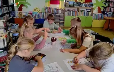 Dzieci przy białym rozłożonym stole w bibliotece kolorują na kartkach smoki.