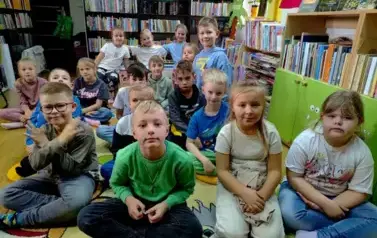 Na dywanie siedzi liczna grupa dzieci słuchająca czytanej bajki.