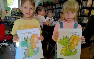 Chłopiec i  dziewczynka w blond włosach pokazują swoje pokolorowane smoki. W tle widać inne dzieci.
