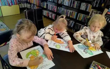 Trzy dziewczynki w blond włosach siedzą przy stole i kolorują starannie smoki.