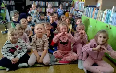 Dzieci liczną grupą siedzą na dywanie, dwie dziewczynki pokazują palcami serduszko.