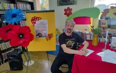 Pan Pilarski siedzi przy czerwonym stole i podpisuje książkę dla czytelnik&oacute;w ze Zblewa. Obok jest tablica z plakatem dot. spotkania i duże kwiaty zrobione z krepy.
