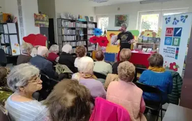 Widok z tyłu sali, krzesła zajęte przez naszych gości. Z przodu Pan Pilarski recytuje wiersze gestykuląc rękami.