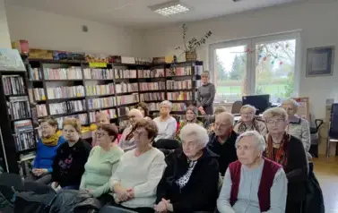 Twarze senior&oacute;w siedzących na krzesłach, a za biurkiem stoi bibliotekarka.