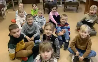 Dzieci siedzą na siedziskach, z tyłu na krześle siedzi Pani z biblioteki z książką w rękach.
