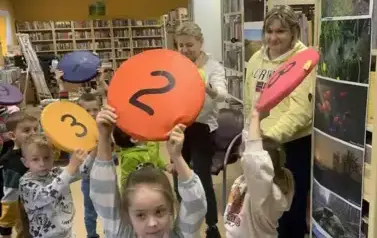 Dzieci stoją w rozsypce i trzymają w g&oacute;rze siedziska z numerkami. Obok stoją ich dwie Panie.
