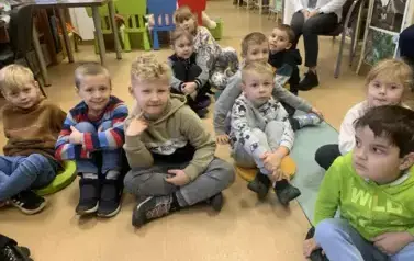 Dzieci siedzą na siedziskach i słuchają bajki. Z tyłu siedzi ich Pani na krześle.