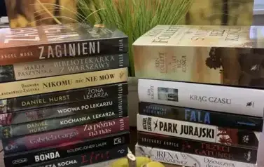 Na stole leżą książki ułożone pionowo, przy nich leżą dynie dekoracyjne. W tle jesienny pejzaż.