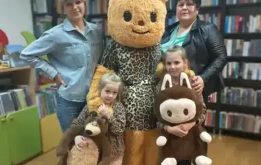 Misia w przebraniu stoi w środku z dwiema dziewczynkami, kt&oacute;re trzymają swoje misie. Po bokach stoją Aneczka Bajeczka i Pani z biblioteki.