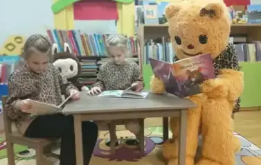 Dwie dziewczynki i Misia w przebraniu siedzą przy małym stoliku w Pinczynie i czytają bajeczki