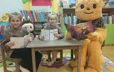 Dwie dziewczynki i Misia w przebraniu siedzą przy małym stoliku w bbiliotece w Pinczynie i mają otwarte książeczki.