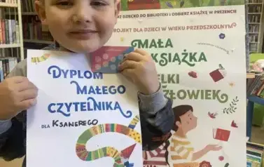 Uśmiechnięty chłopiec pokazuje sw&oacute;j dyplom, za nim stoi tablica z plakatami dot. projektu Mała Książka Wielki Człowiek.