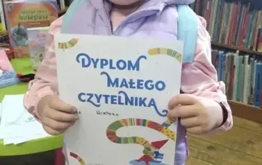 Dziewczynka w siwej opasce na głowie pokazuje sw&oacute;j dyplom w bibliotece w Borzechowie.