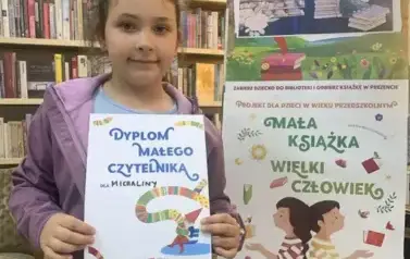 Dziewczynka w ciemnych włosach pokazuje sw&oacute;j dyplom Małego Czytelnika, obok stoi tablica z plakatami dot. projektu Mała Książka Wileki Człowiek