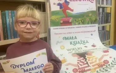 Dziewczynka w blond włosach i okularach trzyma sw&oacute;j dyplom Małego Czytelnika. Obok stoi tablica z plakatami dot. projektu.