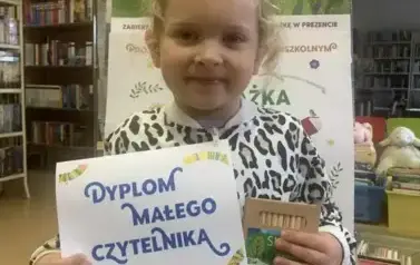 Dziewczynka w lekko kręconych blond włosach pokazuje sw&oacute;j dyplom i kredki świecowe. Za nią stoi tablica z plakatami dotyczącymi projektu Mała Książka Wielki Człowiek.