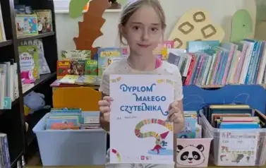 Dziewczynka w blond włosach siedzi na krzesłku w bibliotece w Pinczynie i pokazuje sw&oacute;j dyplom Małego Czytelnika.