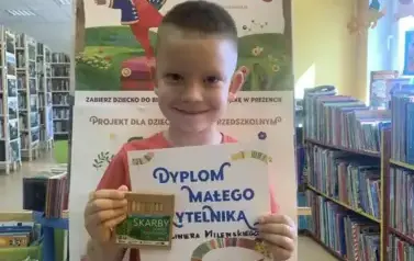 Uśmiechnięty chłopiec pokazuje sw&oacute;j dyplom i kredki. Za nim stoi tablica z plakatami Mała Książka Wielki Człowiek