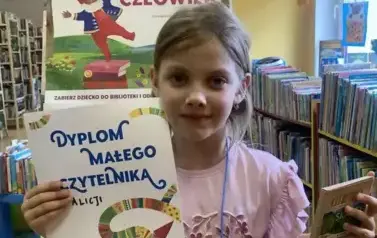Dziewczynka w blond włosach pokazuje sw&oacute;j dyplom i kredki świecowe. Za nią stoi tablica z plakatami Mała Książka Wielki Człowiek.