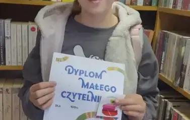 Dziewczynka w upiętych blond włosach pokazuje sw&oacute;j dyplom w bibliotece w Borzechowie.