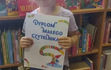 Dziewczynka w blond włosach upiętych stoi przy regałach w bibliotece w Borzechowie i trzyma dyplom Małego Czytelnika.