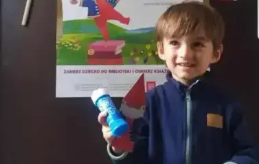 Uśmiechnięty chłopiec w kasztanowych włosach pokazuje sw&oacute;j dyplom, zakładkę do ksiązki i bańki mydlane w pojemniczku. Stoi przy drzwiach na kt&oacute;rych wisi plakat Mała Książka Wielki Człowiek