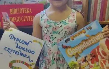 Dziewczynka w blond wlosach w kolorowej sukience pokazuje sw&oacute;j dyplom i wypożyczoną książeczkę w bibliotece w Borzechowie.