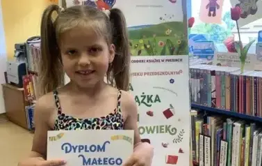 Dziewczynka w blond włosach uczesanych w dwa kitki pokazuje sw&oacute;j dyplom Małego Czytelnika. Za nią stoi tablica z plakatami Mała Książka Wielki Człowiek