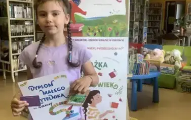 Dziewczynka w ciemnych warkoczach trzyma dyplom, kredki i kartę Małego Czytelnika. Za nią stoi tablica z plakatami dotyczącymi projektu Mała Książka Wielki Człowiek