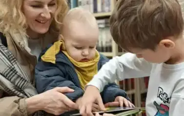 Matka siedzi z dzieckiem na kolanach,a przy nich stoi chłopiec i przeglądają książeczkę.
