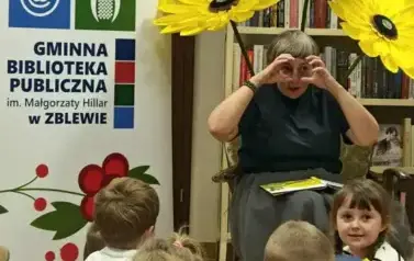 Dzieci siedzą na siedziskach i się rozglądają wokoło. Z przodu Pani Małgosia ma położone książkę na kolanach, a dłonie ułożone przy oczach w kształcie okular&oacute;w. Za Nią stoją trzy sztuczne kwiaty z krepy, po lewej rolap z logiem biblioteki.