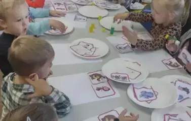 Dzieci siedzą przy białym stole w Borzechowie i na talerzykach mają części układanki  z postaciami z bajek i na białej kartce przyklejają pasujace części bohater&oacute;w z bajki. Ich Pani pochylona jest nad stołem trzymając klej.