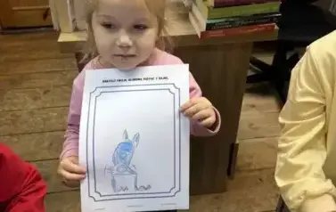 Mała dziewczynka w blond włosach pokazuje swoją narysowaną postać z bajki.