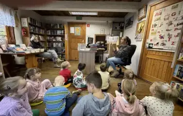 Dzieci siedzą na siedziskach w bibliotece i patrzą w kierunku bibliotekarki, kt&oacute;ra czyta im bajkę pokazując przy tym ilustracje. Z lewej strony przy stole siedzi Pani z Przedszkola.
